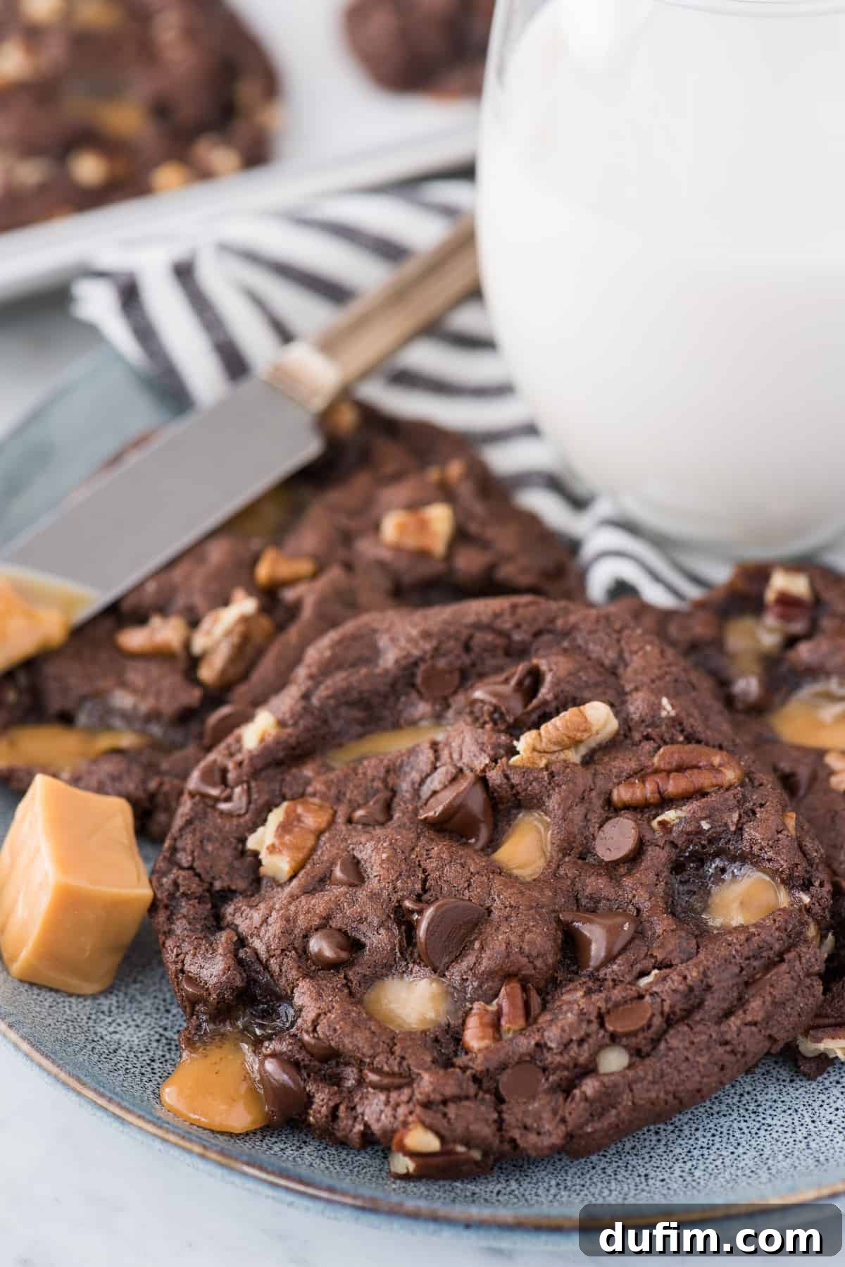 Delicious chocolate turtle cookies, loaded with caramel, pecans, and chocolate chips, arranged artfully on a blue plate with a refreshing glass of milk in the background, ready to be enjoyed.