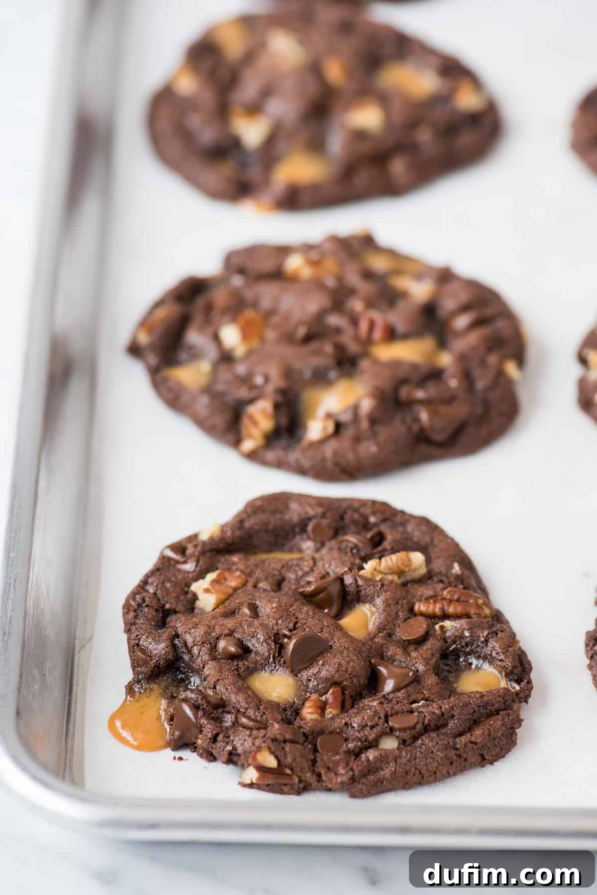 Batch of freshly baked chocolate turtle cookies featuring visible caramel swirls, chopped pecans, and melted chocolate chips, cooling on white parchment paper on a metal baking sheet.