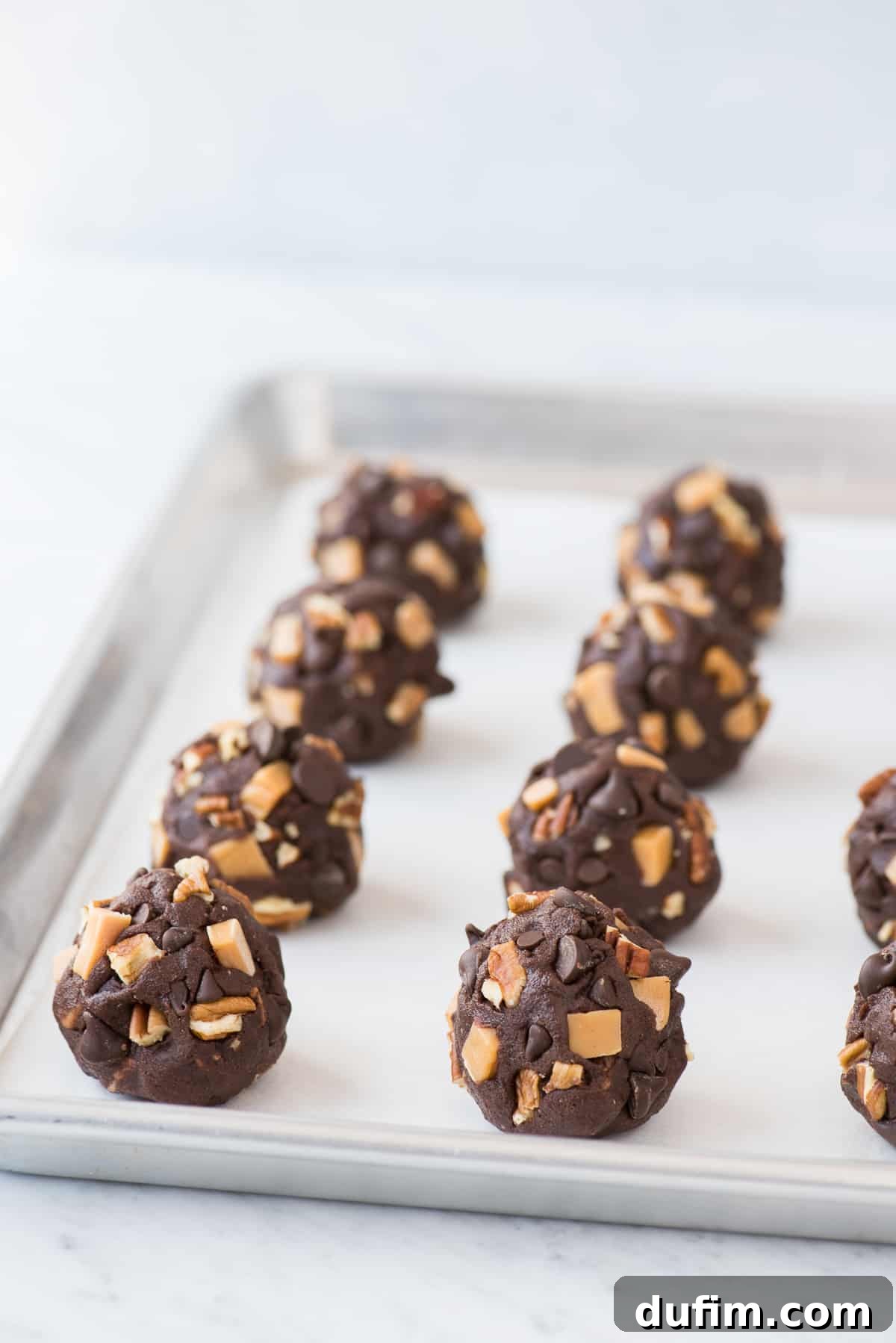 Portion-controlled chocolate turtle cookie dough balls, generously adorned with caramel pieces, chopped pecans, and chocolate chips, meticulously arranged on white parchment paper on a metal baking sheet, ready for baking.