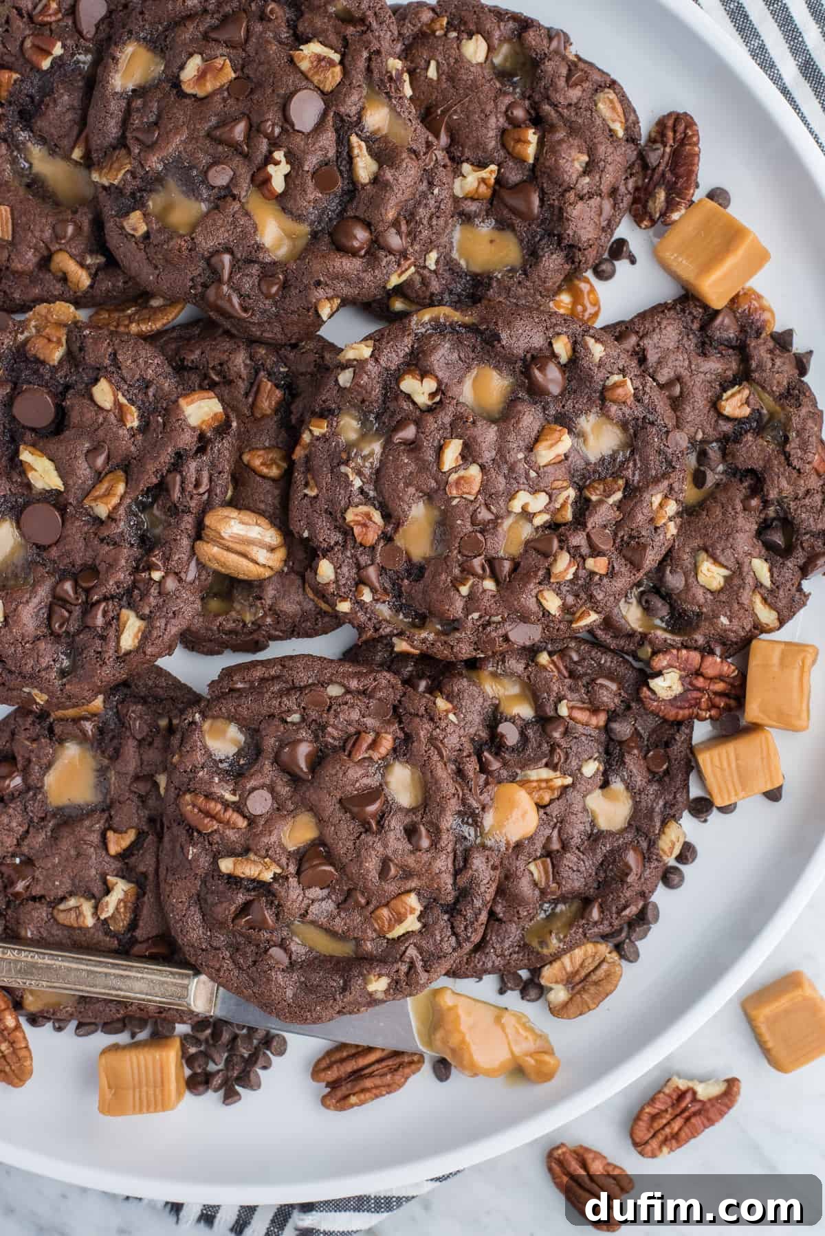 An inviting display of chocolate turtle cookies, generously dotted with caramel, pecans, and chocolate chips, artfully arranged on a white round serving tray, ready to be shared.