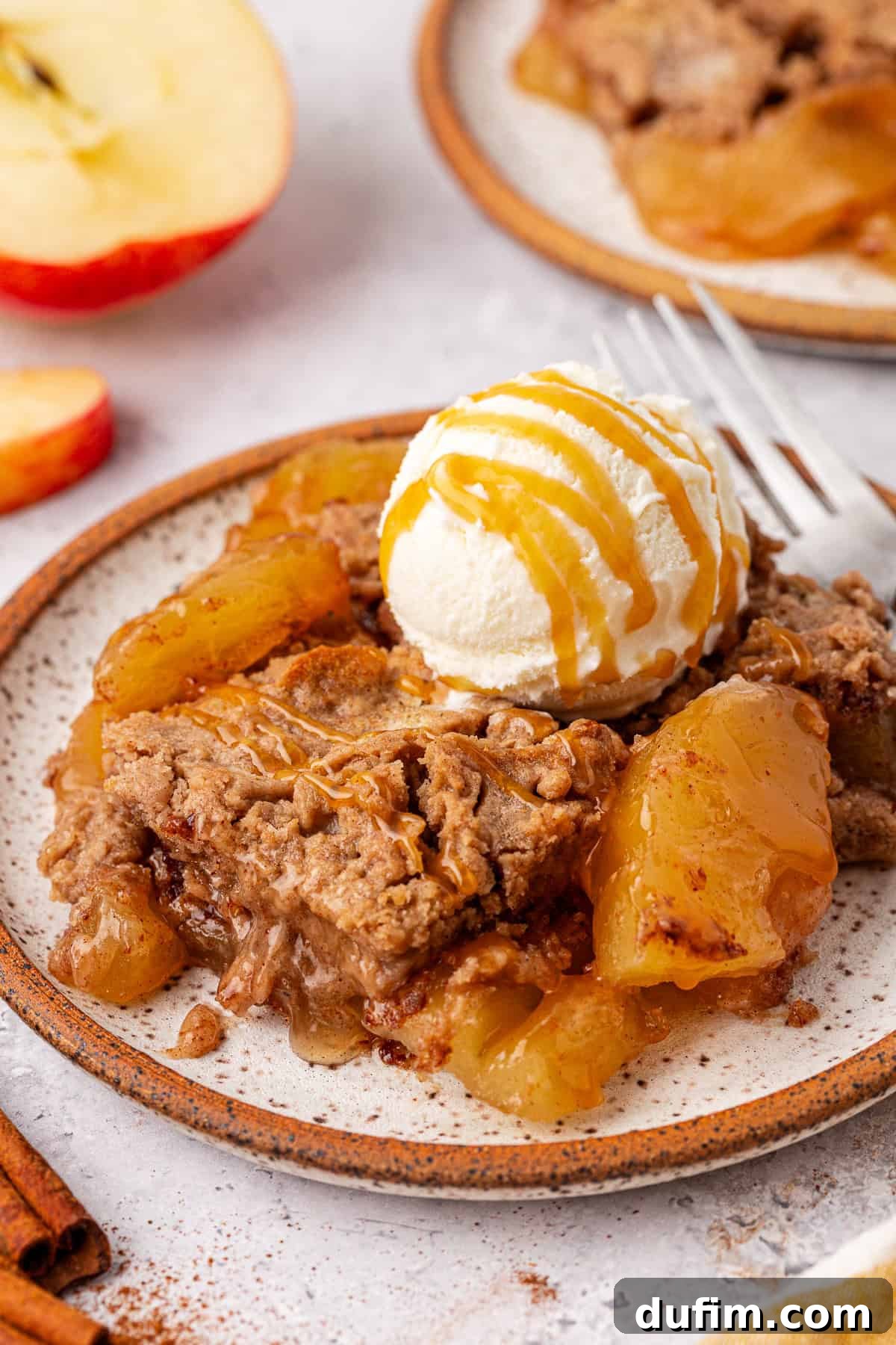 A dessert plate showcasing a warm apple dump cake crowned with a scoop of vanilla ice cream, drizzled in caramel sauce. Slices of apple and a cinnamon stick are artfully arranged in the background on a speckled ceramic plate.