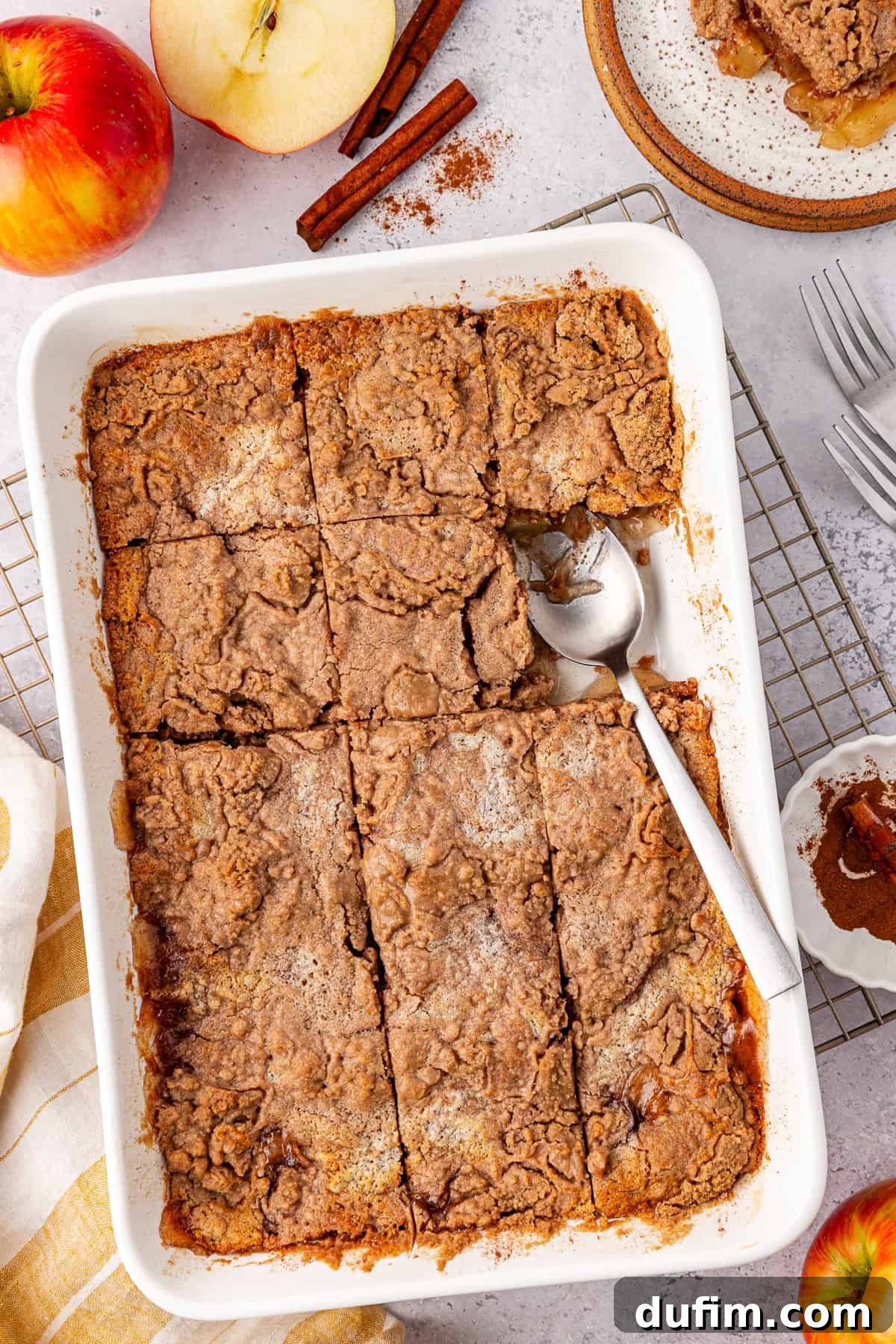 A top view of a baked apple crisp, reminiscent of an apple dump cake, in a rectangular white dish, partially served with a metal spoon. Fresh apples, cinnamon sticks, a plate with a portion of crisp, and a small bowl of cinnamon lie around on the light surface.