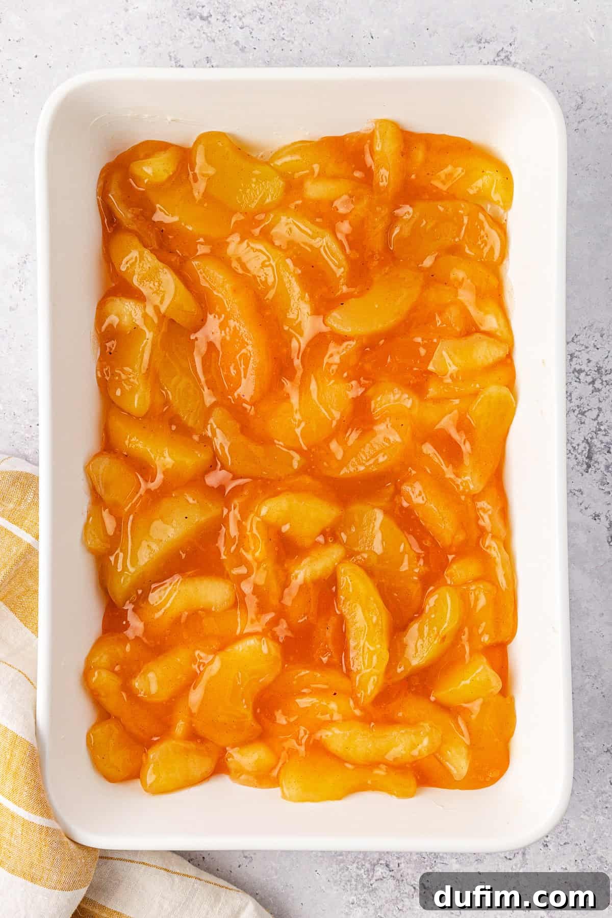 A white baking dish filled with glossy, golden-brown apple pie filling. A folded striped yellow and white cloth napkin is partially visible on the bottom left corner, resting against a light gray surface background.
