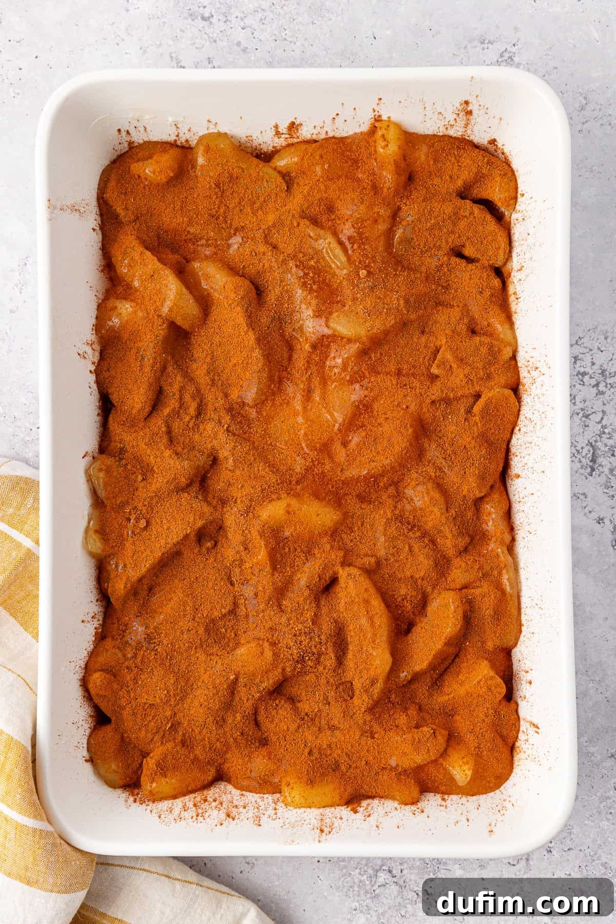 A rectangular baking dish brimming with apple dump cake features sliced apples topped abundantly with cinnamon and sugar, forming a caramelized crust. The dish rests on a light gray surface beside a white and yellow striped cloth.