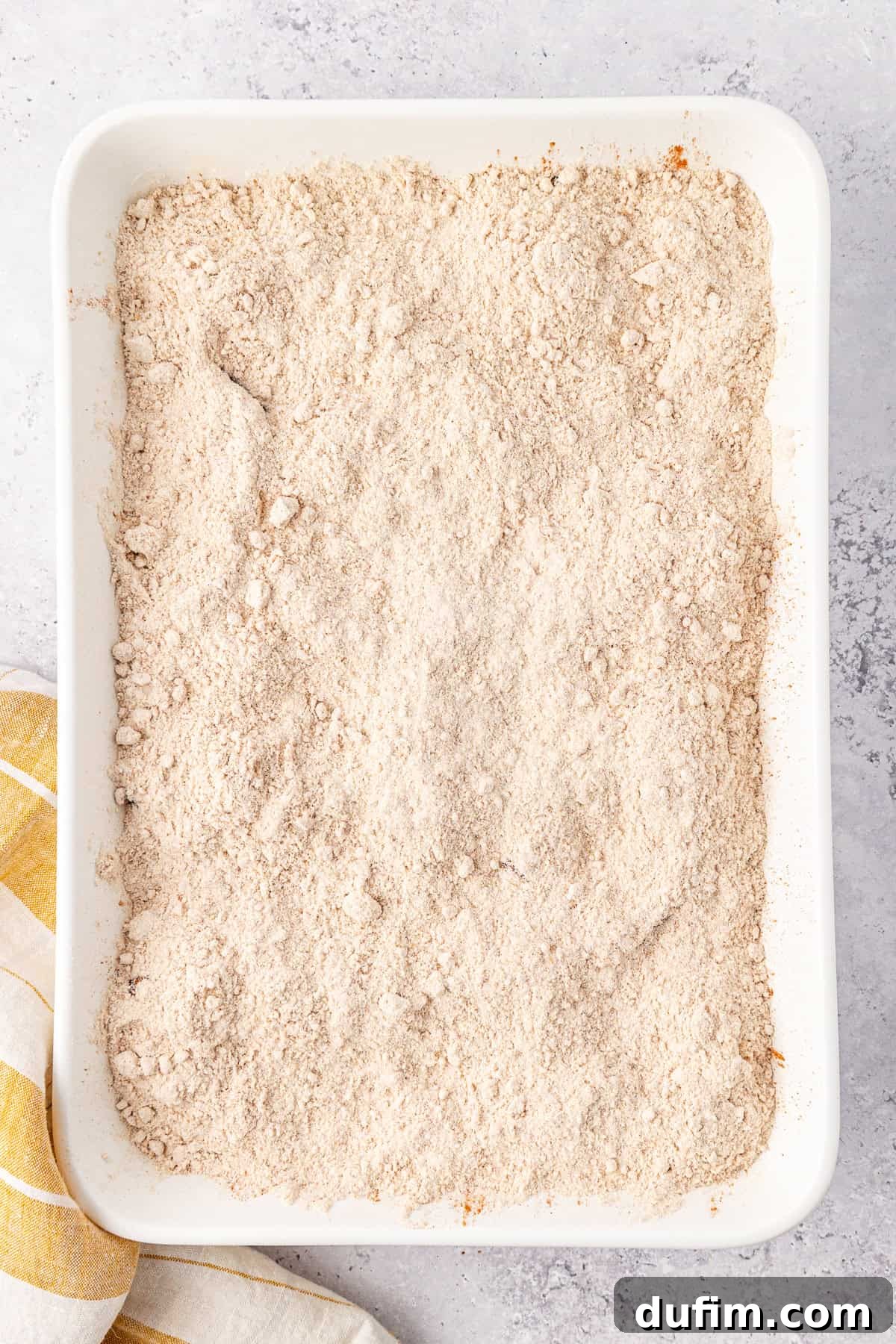 A rectangular white baking dish filled with an even layer of cake mix topping for an apple dump cake, sits on a light gray surface. A yellow and white striped cloth is partially visible beside the dish.