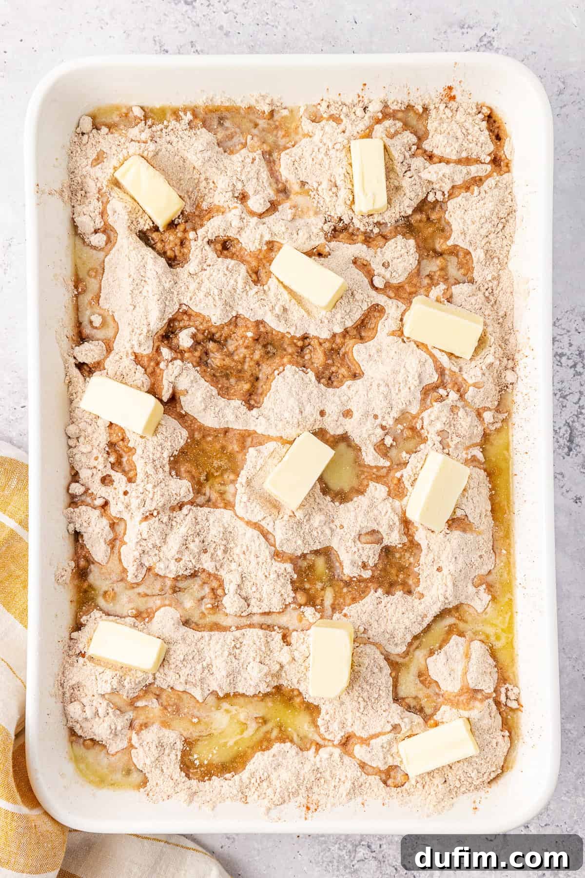 Rectangular baking dish with a apple dump cake before baking. Slices of butter are scattered atop dry cake mix and peaches, reminiscent of an apple dump cake. The dish rests on a textured surface, with a yellow and white cloth draped nearby.