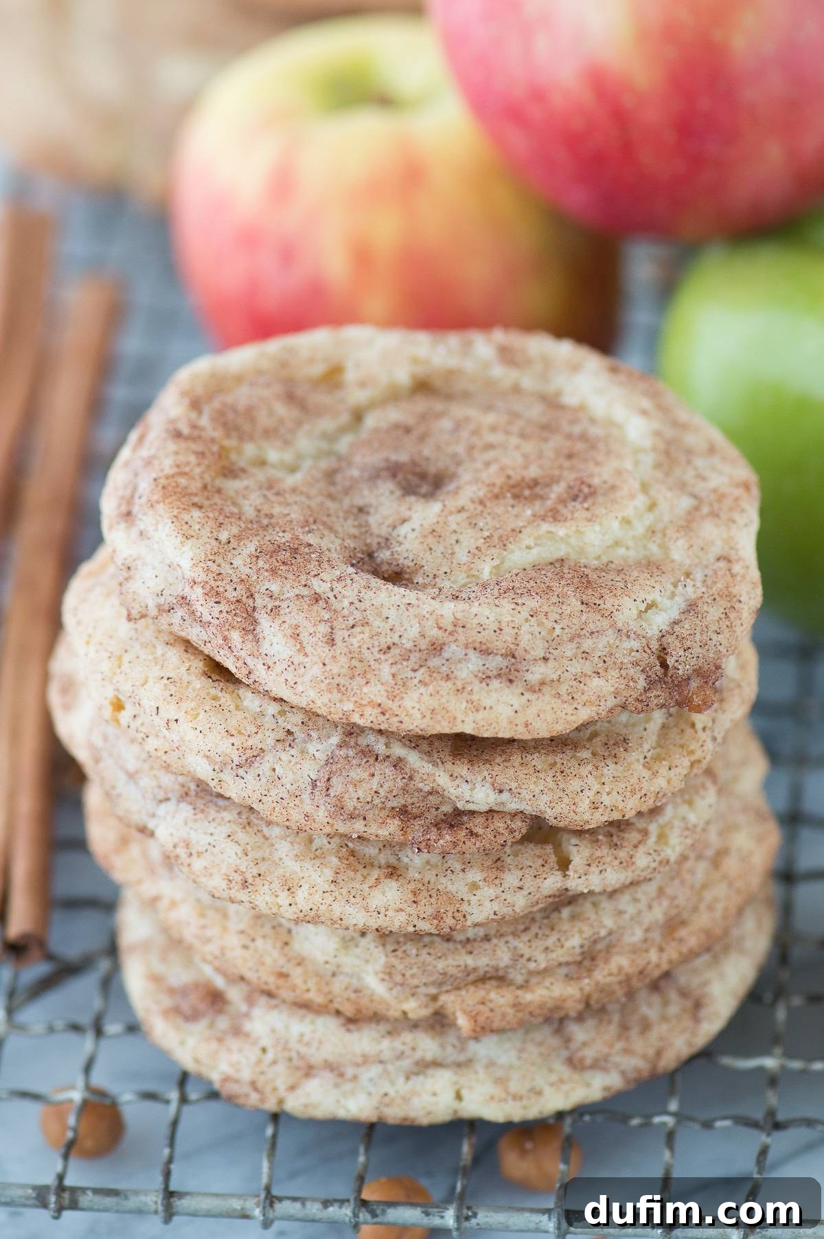 Stacked caramel apple snickerdoodle cookies on a metal cooling rack with fresh apples in the background, hinting at autumn's bounty.