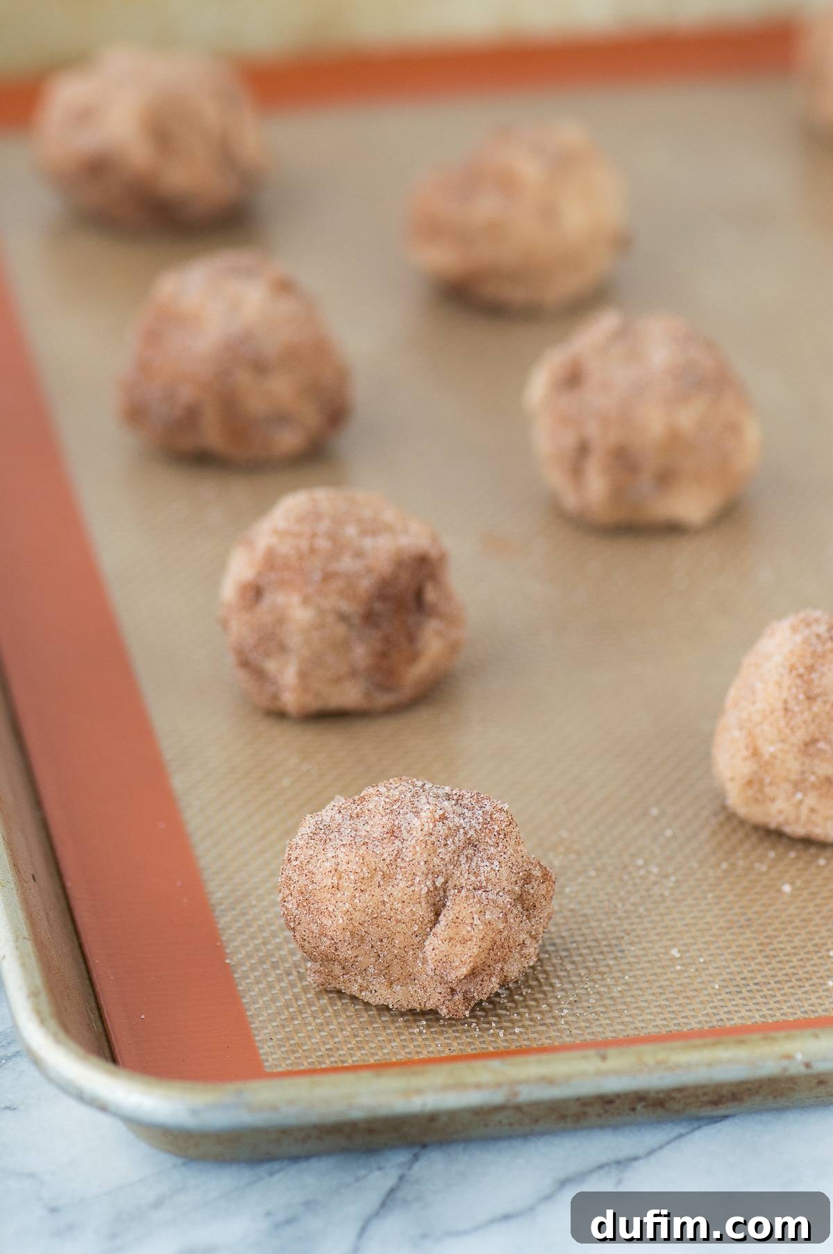Neatly arranged caramel apple snickerdoodle dough balls on a silicone baking mat atop a metal baking sheet, ready for the oven.