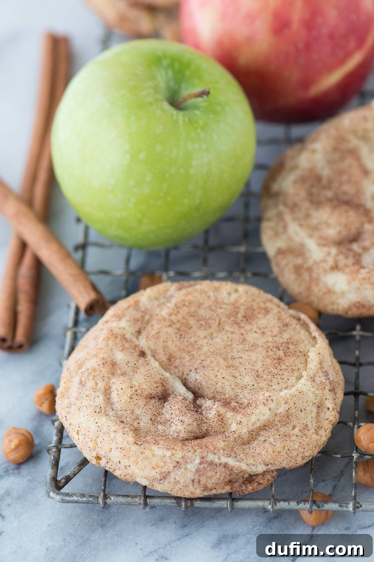 A single caramel apple snickerdoodle cookie resting on a wire cooling rack, with scattered caramel bits, cinnamon sticks, and a fresh apple creating a rustic fall ambiance in the background.