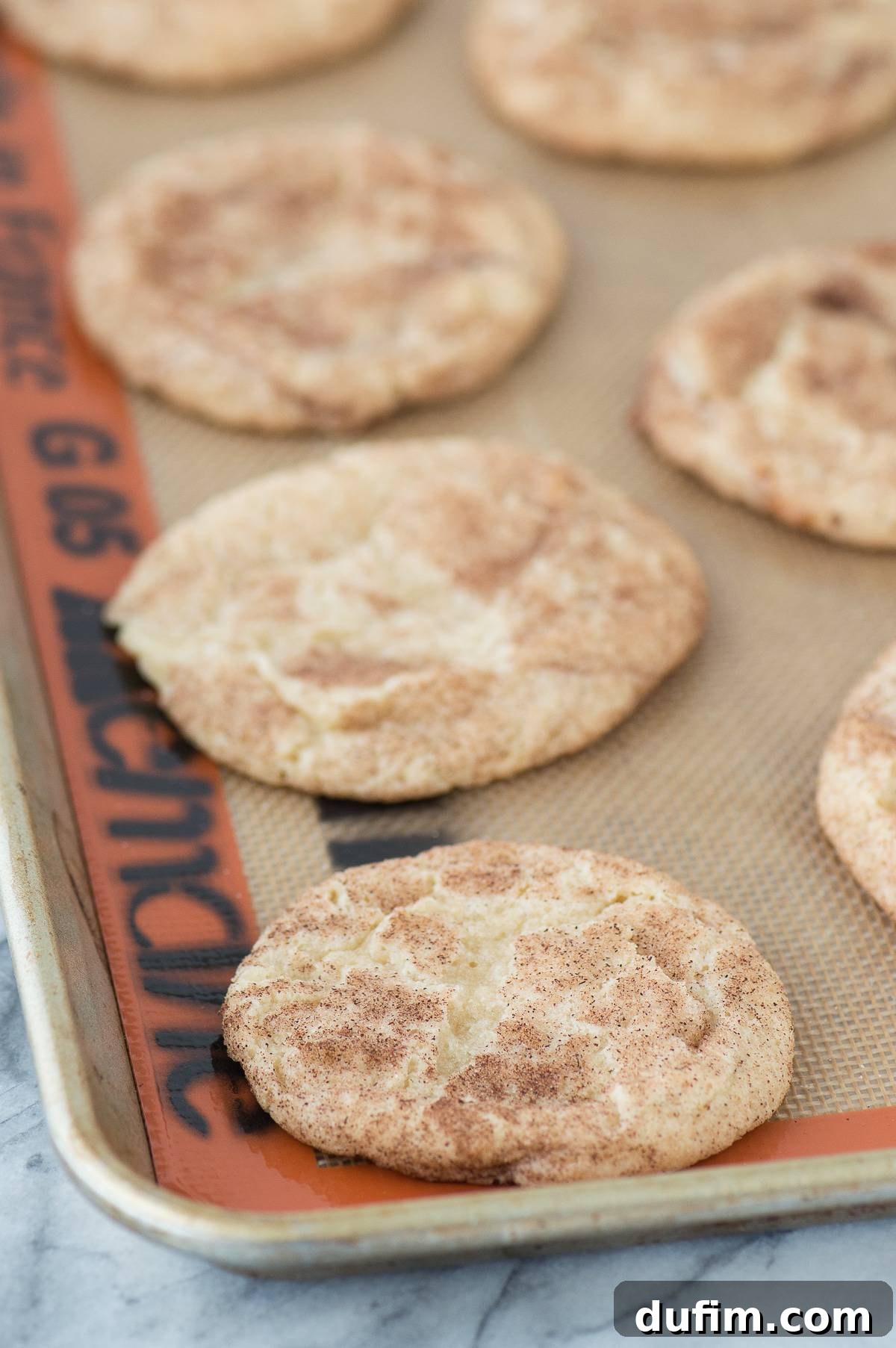 Rows of perfectly baked Caramel Apple Snickerdoodles cooling on a silicone baking mat on a metal sheet, showcasing their golden color and crackled tops.