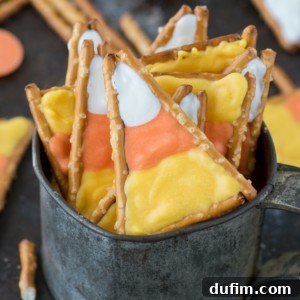 A metal cup filled with homemade candy corn pretzels, showcasing their vibrant white, orange, and yellow layers. Festive Halloween snacks are blurred in the background.