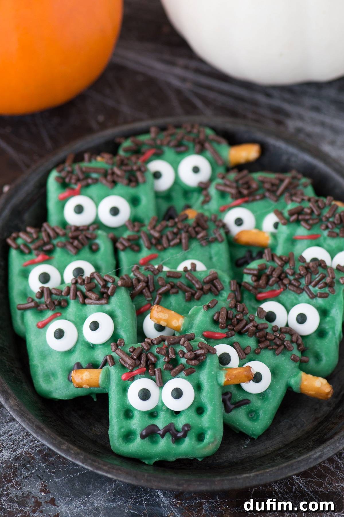 Green Frankenstein treats made from square waffle pretzels, dipped in green chocolate with decorative embellishments, presented on a dark plate against a dark background, showcasing their spooky charm.