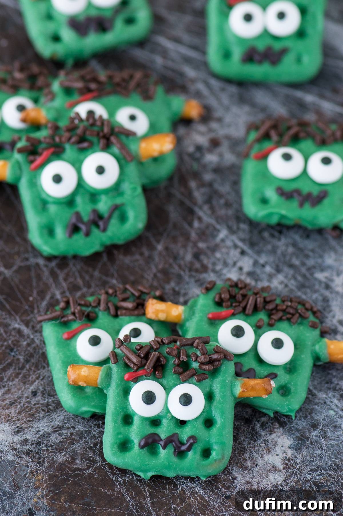 Individual Frankenstein treats made from square waffle pretzels dipped in green chocolate, intricately decorated with candy eyes, sprinkle hair, and pretzel bolts, presented on a dark background.