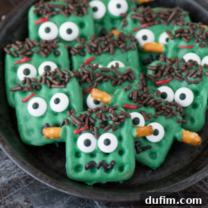 Green, rectangular cookies decorated as Frankenstein’s monster with candy eyes, chocolate sprinkle hair, red icing scars, and pretzel bolts are perfect for a playful Frankenstein Halloween treat. Each cookie has a fun, expressive face in a dark bowl.