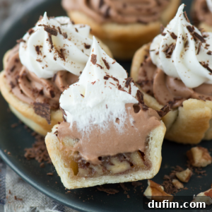 Mini chocolate silk pecan pies topped with swirls of chocolate and whipped cream, garnished with chocolate shavings, are arranged on a plate. One pie is cut open, revealing a nutty filling inside the flaky crust.