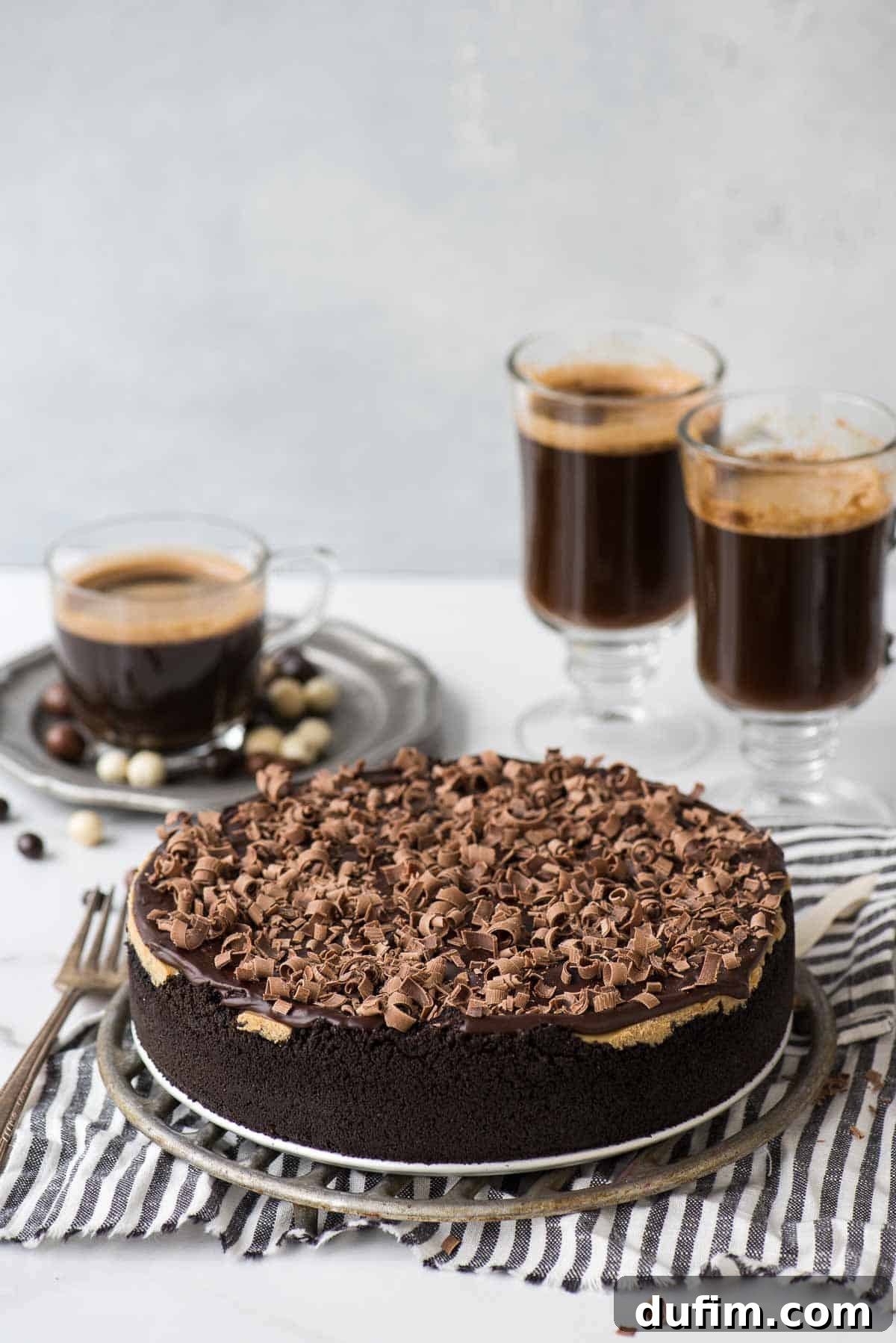 Decadent espresso cheesecake slice on a metal stand with coffee cups in the background, showcasing its rich texture and topping.
