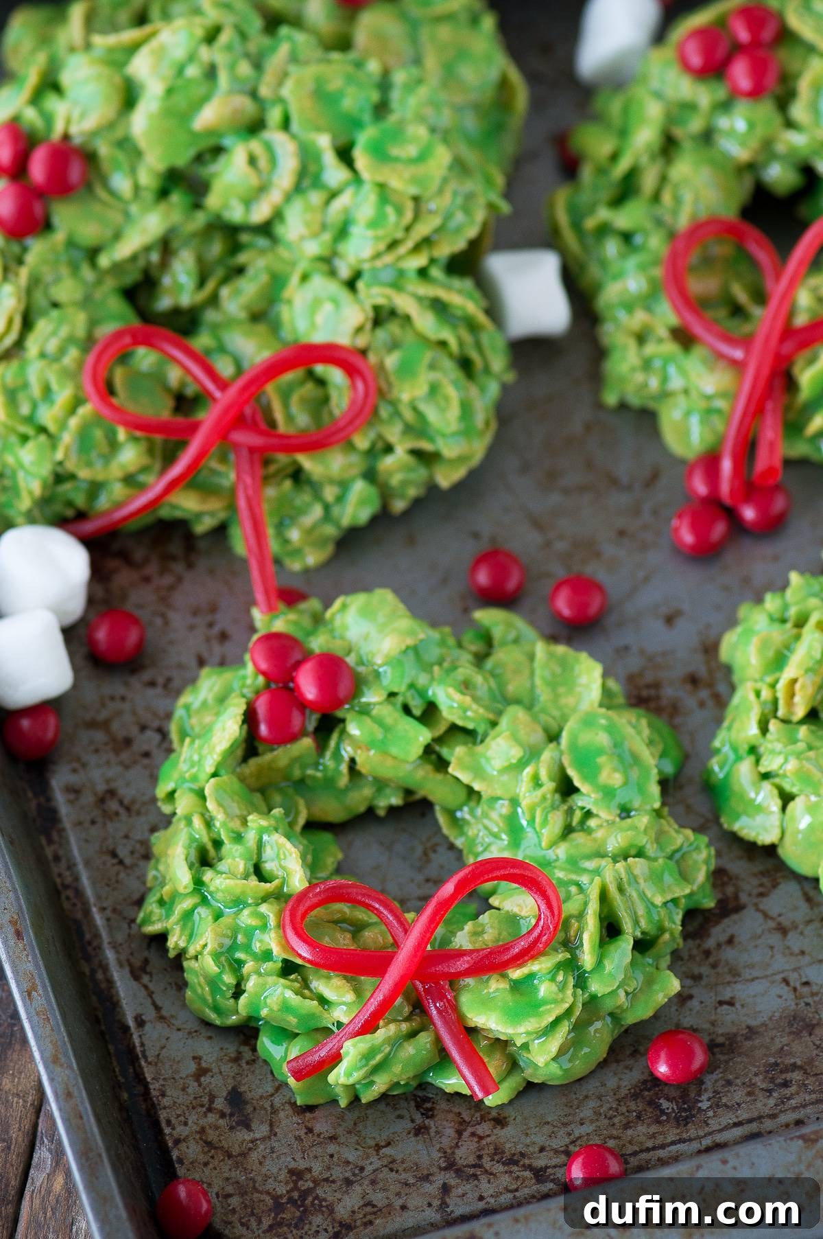 Golden No-Bake Wonders 2 Vibrant green Christmas wreath cookies made with cornflakes, adorned with an edible red licorice bow, resting on a rustic metal surface, showcasing their festive appeal.