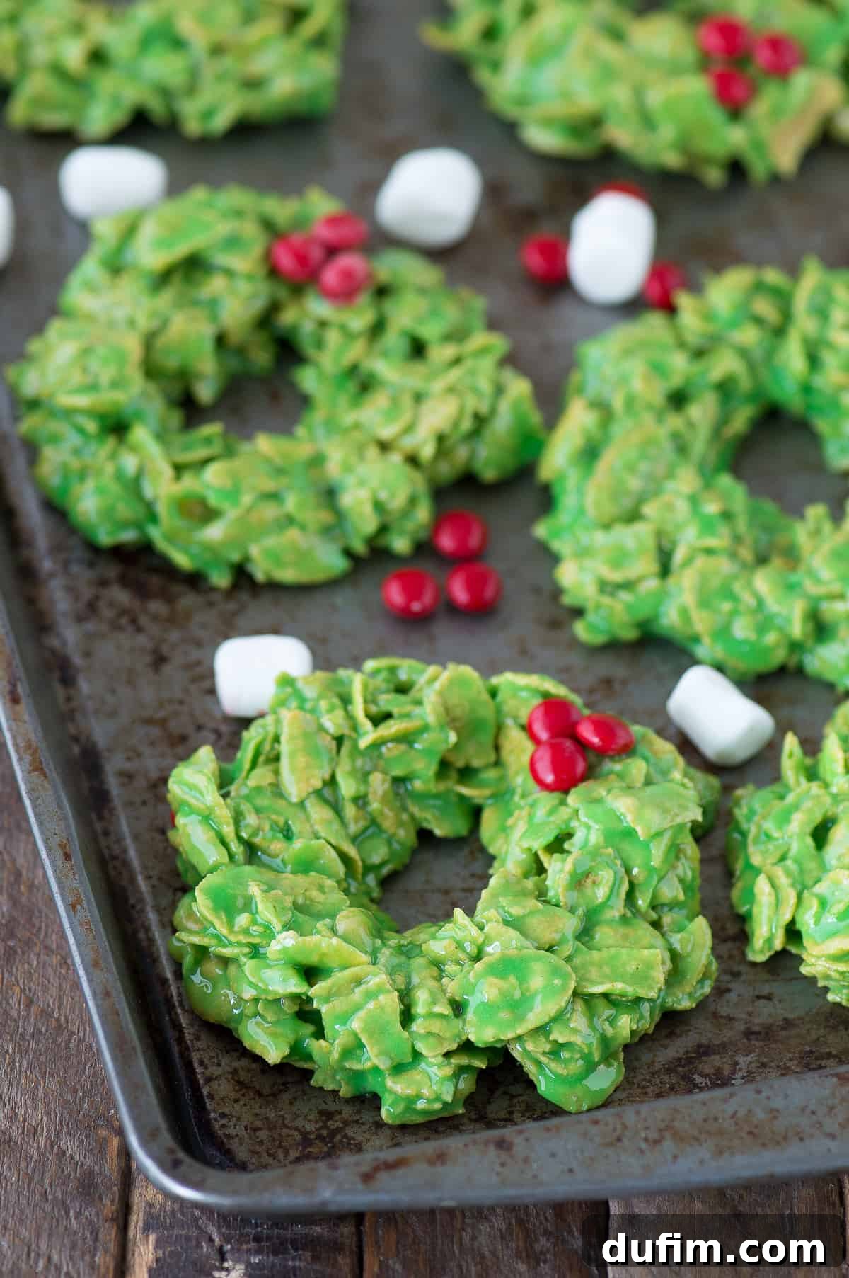 Golden No-Bake Wonders 11 A beautifully shaped green cornflake wreath cookie, perfectly adorned with vibrant red candies and a delicate licorice bow, presented on a sleek metal surface.