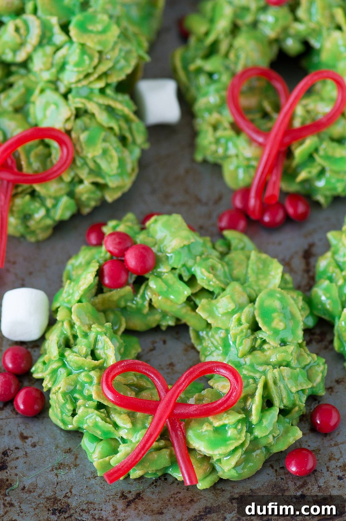 Golden No-Bake Wonders 13 Close-up of a festive green cornflake wreath cookie, decorated with a bright red edible bow, perfectly showcasing its holiday charm on a metal surface.