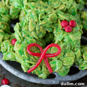 Golden No-Bake Wonders 14 Close-up of a festive green cornflake wreath cookie decorated with a licorice bow and vibrant red candy berries, presented on a rustic metal tray.