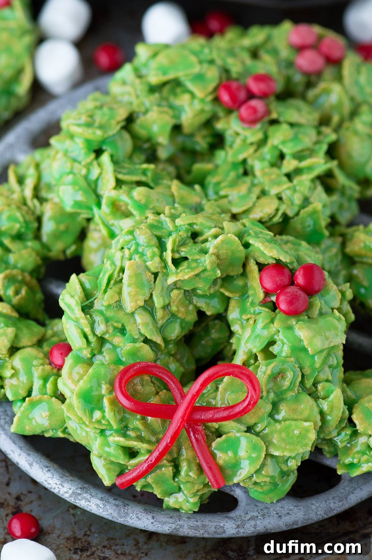 Golden No-Bake Wonders 3 Close-up of three festive green Christmas wreath cookies, decorated with red candies and licorice bows, arranged artfully on a metal surface, ready for a holiday gathering.