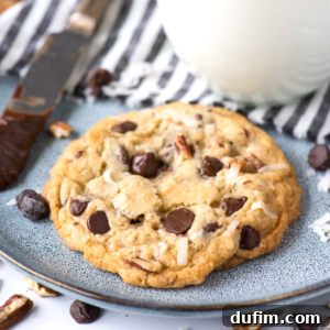 A beautifully arranged Cowboy Cookie with chocolate chips, shredded coconut, and nuts, presented on a gray plate with a knife and glass of milk in the background.