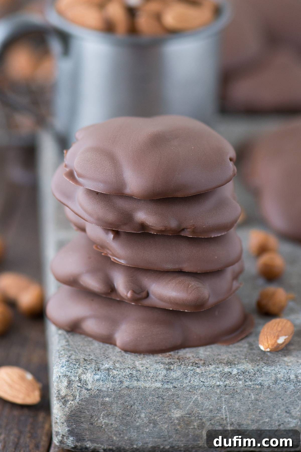 Sticky Paws Williams Sonoma Copycat Christmas candy recipe! Almond caramel clusters dipped in chocolate. Make these at home for a fraction of the price! A stack of five gourmet Sticky Paws next to a scattering of whole almonds on a rustic wooden board, highlighting the fresh ingredients.