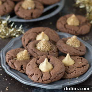 A plate of midnight kiss cookies, topped with gold-dusted chocolate kisses, is shown on a dark surface, surrounded by festive gold decorations and more cookies in the background.