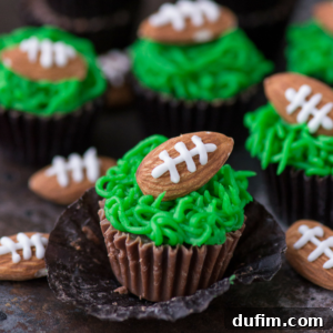 Chocolate cupcakes with green frosting resembling grass, topped with oval cookies decorated like footballs using white icing, perfect for serving as football cups at your next game day party.