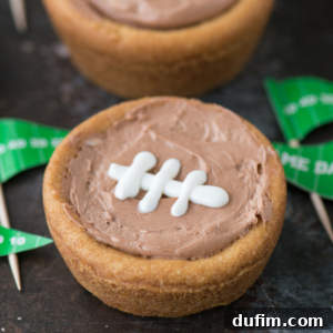 A sugar cookie football cup filled with light brown frosting, decorated with white icing in the shape of football laces, sits on a dark surface with green football-themed paper decorations nearby.