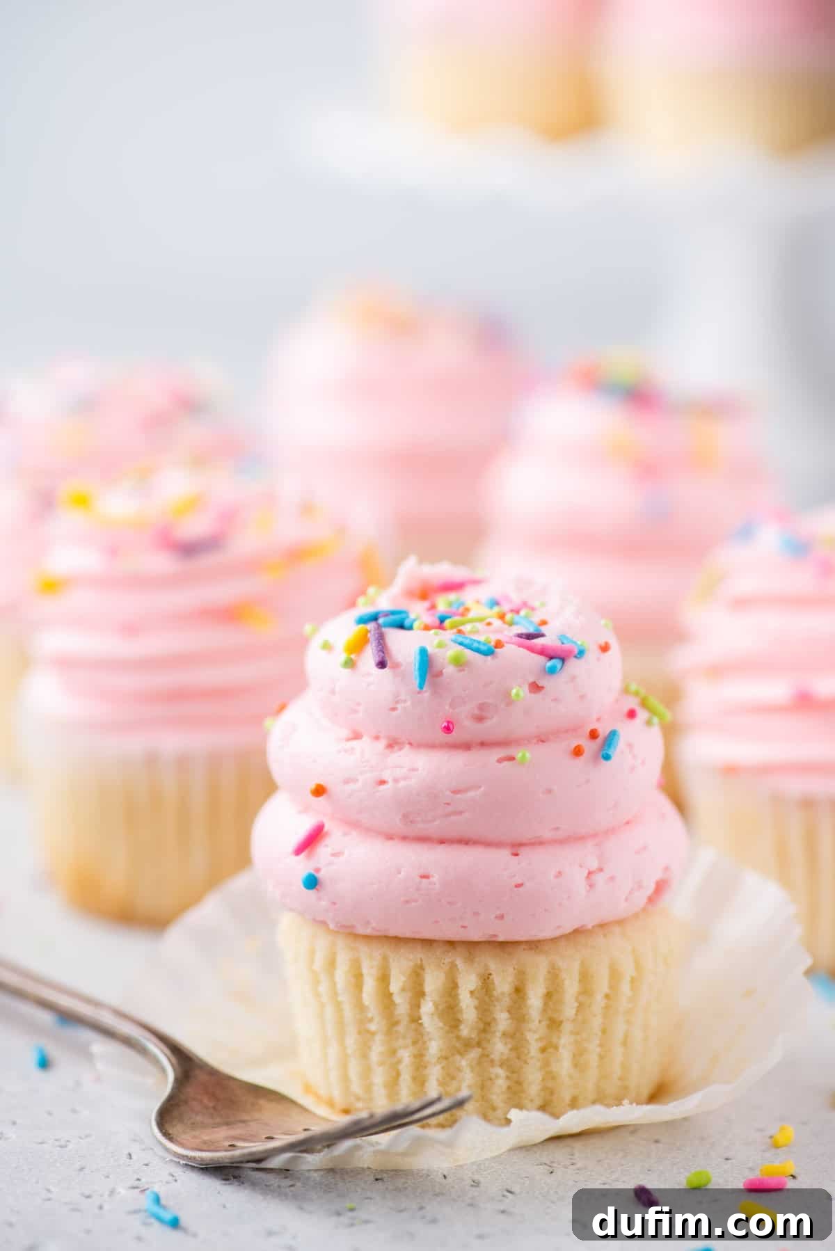A beautifully frosted vanilla cupcake with delicate pink frosting and colorful sprinkles, with its paper liner removed, next to a silver fork. More cupcakes are blurred in the background, hinting at a larger batch.