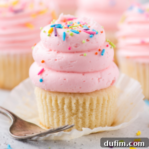 A white cupcake with pink swirled frosting and colorful sprinkles sits on a white wrapper next to a silver fork, with more vanilla cupcakes blurred in the background.