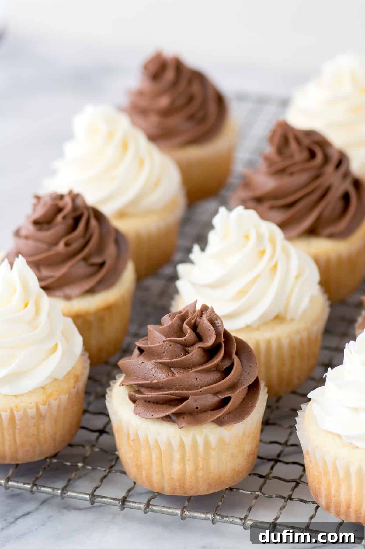 A collection of freshly baked vanilla cupcakes, some topped with rich chocolate frosting and others with classic white vanilla frosting, arranged on a metal cooling rack against a stylish marble background.