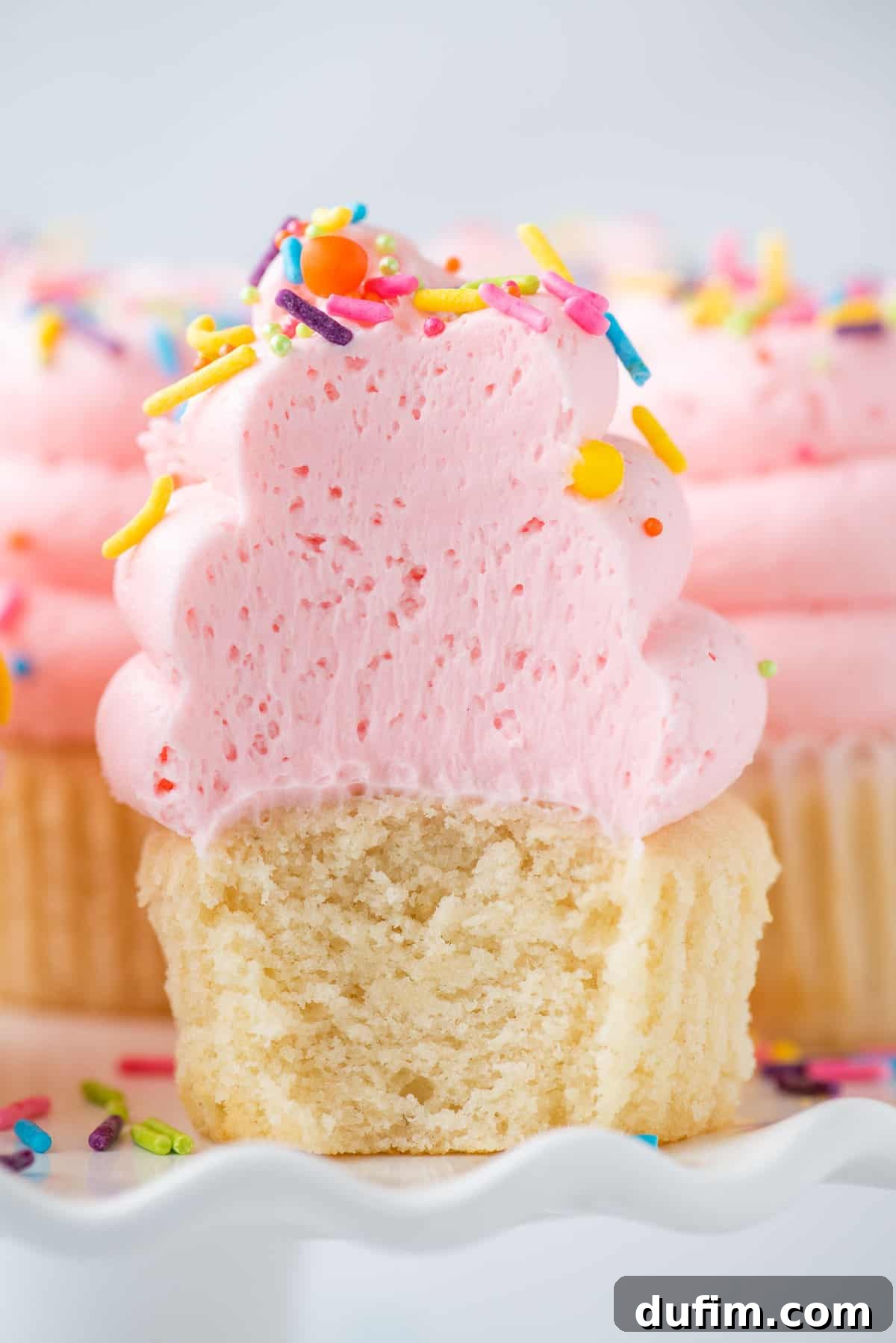 A perfectly baked and frosted vanilla cupcake with pink sprinkles, showing a bite removed to reveal its moist and fluffy white interior.