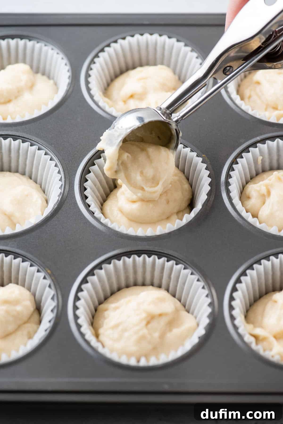 Vanilla cupcake batter being carefully scooped into a muffin pan lined with paper wrappers, ready for baking.