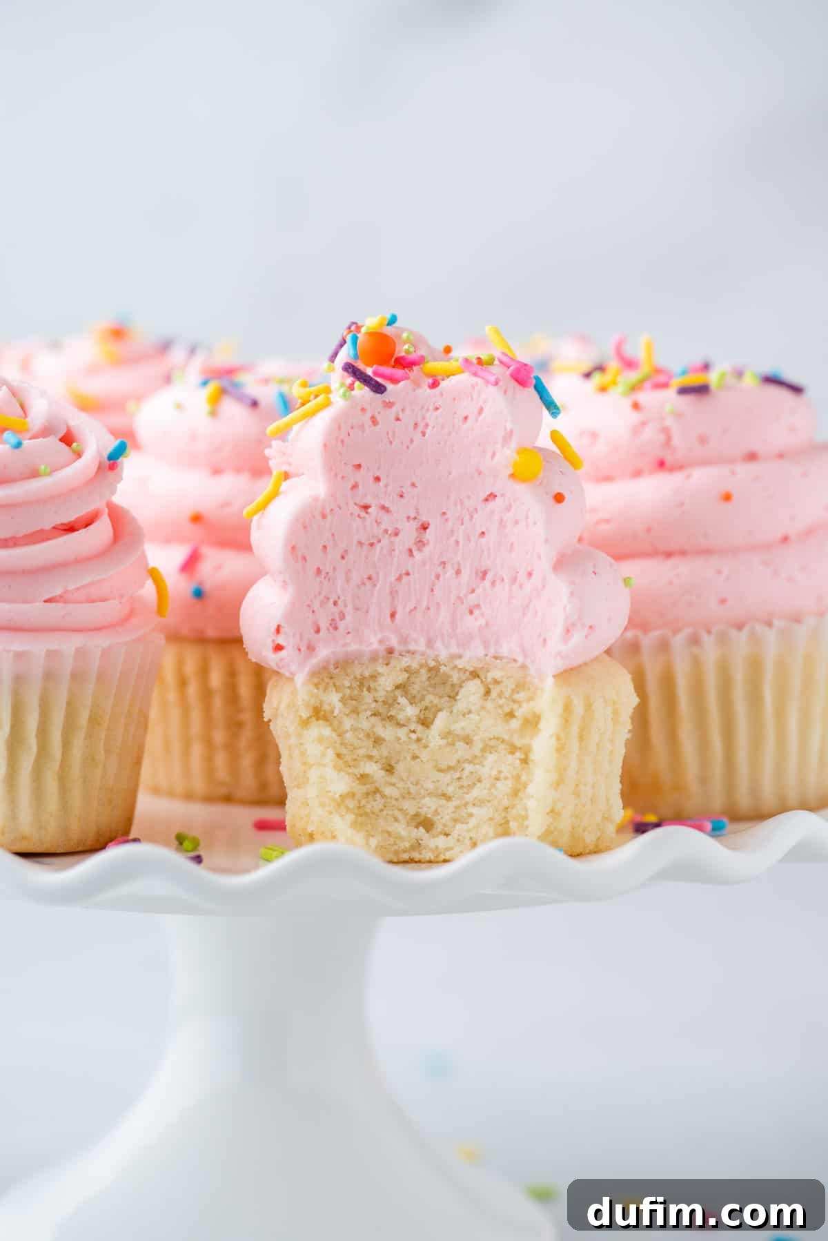 A vanilla cupcake with a generous swirl of pink frosting and colorful sprinkles, resting on a white cake stand, with a bite playfully removed to showcase its perfect texture.