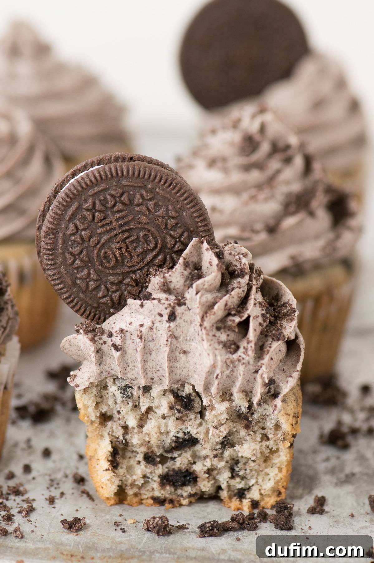 A delicious homemade Oreo cupcake with a bite taken out, showing the moist cake and creamy frosting, resting on a metal baking sheet amidst other cupcakes.