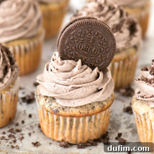 A beautifully decorated Oreo cupcake with swirled cookies and cream frosting, topped with an Oreo cookie, surrounded by other similar cupcakes and cookie crumbs.