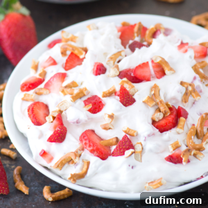 A bowl of creamy dessert topped with sliced strawberries and broken pretzel pieces, with a whole strawberry and more pretzels scattered nearby on a dark surface.