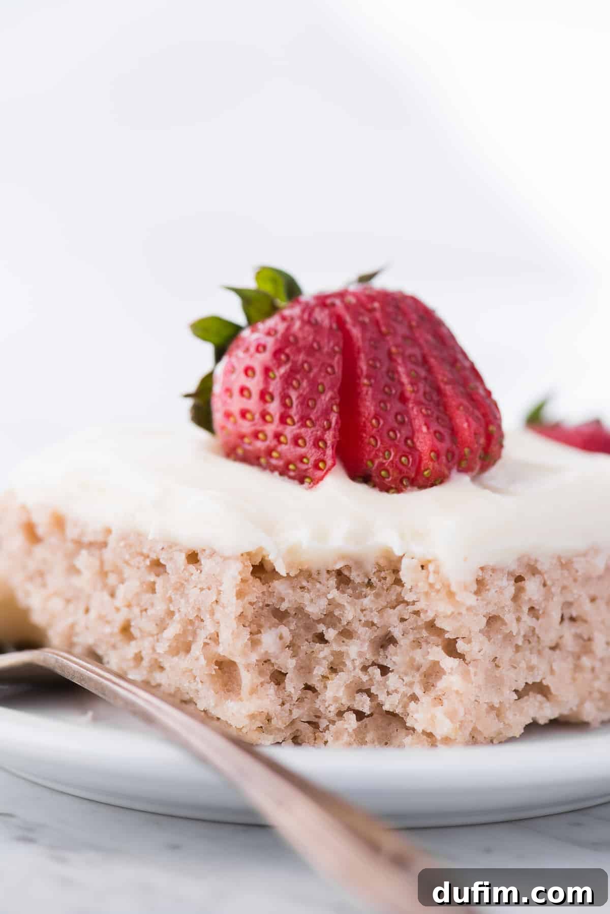 A beautifully frosted strawberry sheet cake with a slice removed, revealing the soft pink crumb, topped with fresh strawberry slices and served on a white plate.