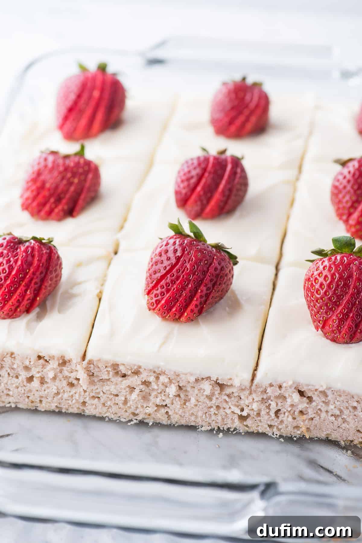 A vibrant strawberry cake with a generous layer of white cream cheese frosting, garnished with fresh strawberry slices, presented in a clear glass baking pan.