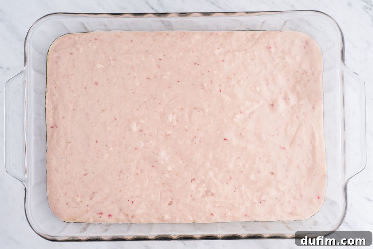 A close-up of light pink strawberry cake batter evenly spread in a clear glass 9x13 inch baking pan, ready for the oven.