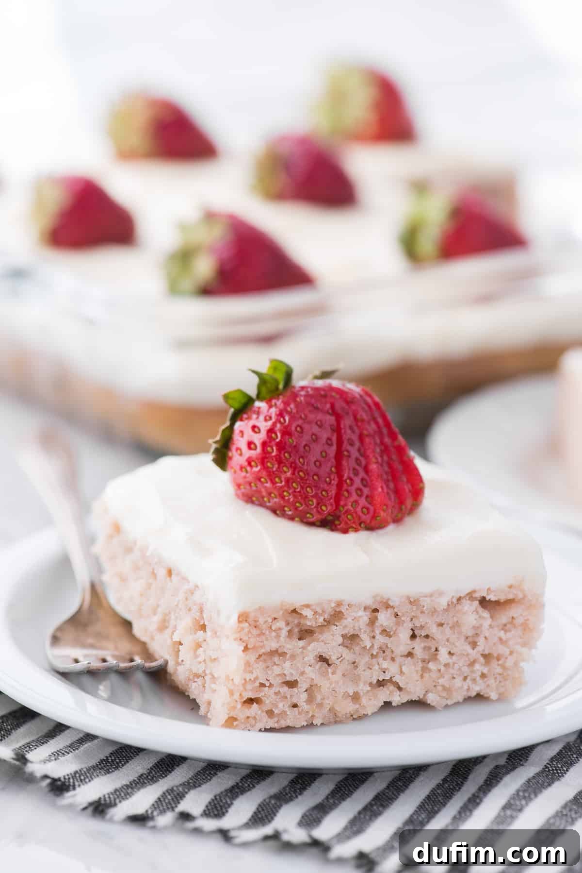 A slice of homemade strawberry cake with white cream cheese frosting and a fresh strawberry on top, served on a small white plate.