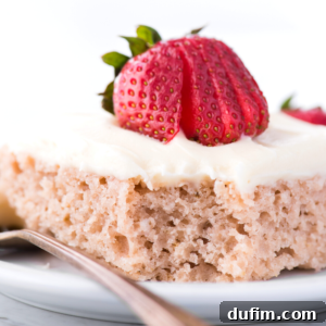 A close-up of a slice of light, fluffy 9x13 inch strawberry cake topped with creamy white frosting and a sliced fresh strawberry, served on a white plate with a silver spoon.
