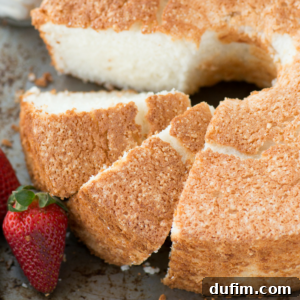 Close-up of sliced angel food cake with a golden-brown crust, showing its light, fluffy texture inside. Two fresh strawberries are placed next to the angel food cake on the plate.