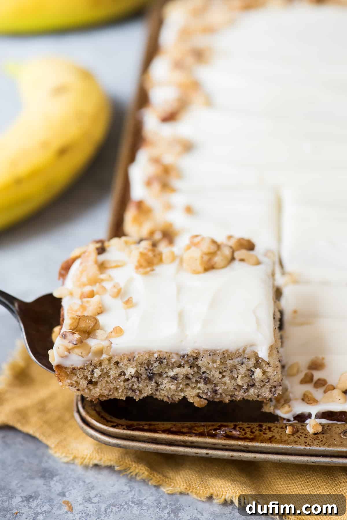 Chewy Banana Nut Bars 3 A slice of moist banana bar with a generous layer of cream cheese frosting, held by a spatula.