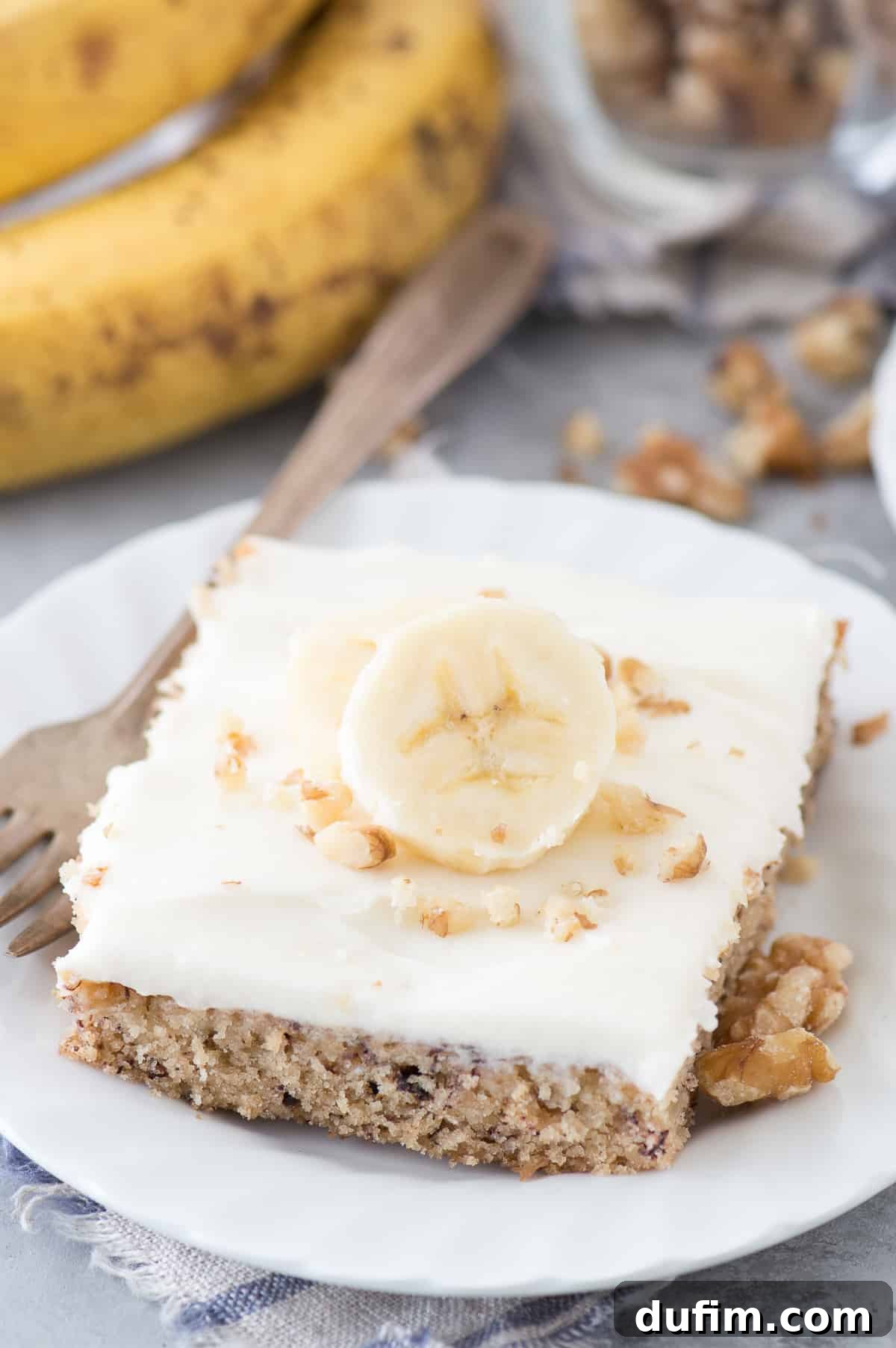 Chewy Banana Nut Bars 6 A single slice of banana bar with cream cheese frosting, garnished with a fresh banana slice, on a white plate.
