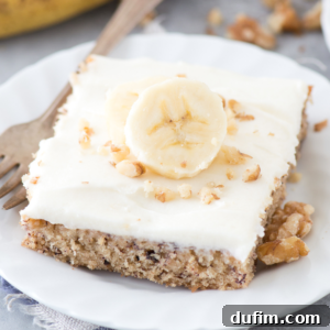 A square slice of banana bars with cream cheese frosting, topped with a banana slice and chopped walnuts, sits on a white plate with a wooden fork beside it.