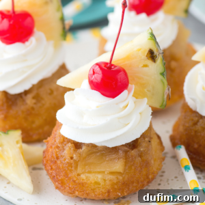 A mini pineapple upside down cupcake topped with a swirl of whipped cream, a maraschino cherry, and a wedge of fresh pineapple, served on a white plate with decorative straws nearby.