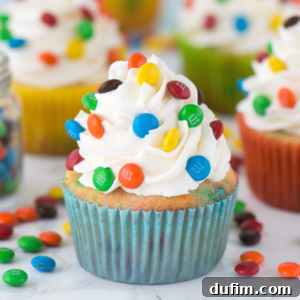 A perfectly decorated M&M cupcake with blue wrapper, white frosting swirls, and colorful M&Ms on top, with more cupcakes and candies in the background.
